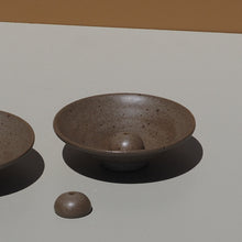 Two grey ceramic incense holders on a light gray surface with a brown background