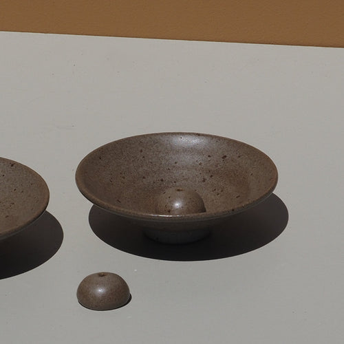 Two grey ceramic incense holders on a light gray surface with a brown background